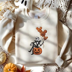 May include: A cream-colored crewneck sweatshirt featuring an embroidered graphic of a skeleton character sitting on a large orange jack-o'-lantern. The character holds three orange balloons shaped like mouse heads with jack-o'-lantern faces.