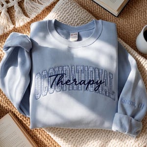 May include: A light blue crewneck sweatshirt featuring the text Occupational Therapy in a blue and white gingham block font, with the word Therapy in dark blue cursive script. The right sleeve is embroidered with the text Emma, OT.