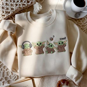 May include: Cream-colored sweatshirt featuring a graphic of four Grogu characters in various poses. One Grogu wears headphones, another has a question mark above its head, and the last one says "cool!".