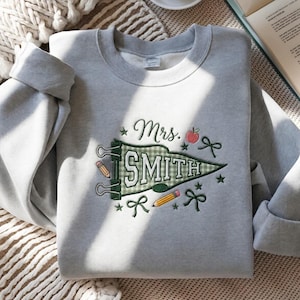 Könnte beinhalten: Ein hellgraues Sweatshirt mit Rundhalsausschnitt und einer Stickerei, die einen grün-weiß karierten Wimpel mit dem Text "Mrs. SMITH" zeigt. Das Design umfasst zwei gelbe Bleistifte, einen roten Apfel, dunkelgrüne Schleifen und kleine Sterne.