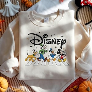 Peut inclure: Un sweat-shirt &agrave; col rond couleur cr&egrave;me arborant un logo Disney brod&eacute; en noir au-dessus d'un motif repr&eacute;sentant Pluto, Dingo, Donald et Mickey marchant sur un passage pi&eacute;ton blanc, agr&eacute;ment&eacute; de petites &eacute;toiles et notes de musique noires.