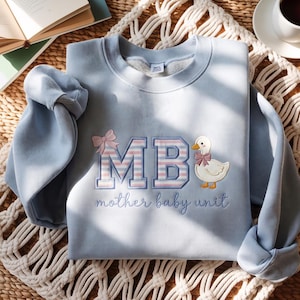 May include: A light blue crewneck sweatshirt featuring embroidered text that reads MB with a pink bow, accompanied by a small white duck graphic wearing a pink bow. Below the letters, the text mother baby unit is embroidered in cursive.