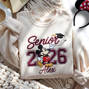 May include: Cream-colored sweatshirt with embroidered "Senior 2026" text and a Mickey Mouse graphic. The design includes a graduation cap and the name "Alex" below the numbers. A Mickey Mouse ear headband with a red bow is also visible.