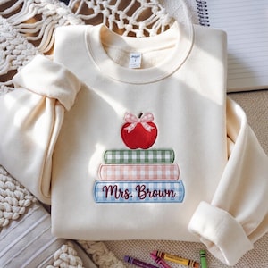 May include: A cream-colored crewneck sweatshirt featuring an embroidered design of three stacked books topped with a red apple wearing a pink gingham bow. The bottom book is light blue with a gingham pattern and the text Mrs. Brown in dark red script.