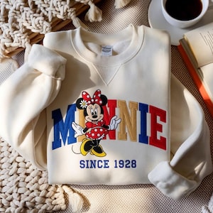 K&ouml;nnte beinhalten: Cremefarbenes Sweatshirt mit Minnie Mouse-Grafik. Das Design zeigt das Wort "MINNIE" in Rot, Blau und Gold, mit Minnie Mouse in einem roten gepunkteten Kleid. Der Text "SINCE 1928" steht darunter.