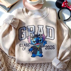 Puede incluir: Sudadera color crema con la palabra "GRAD" en letras grises, y "CLASS OF 2026" debajo. Un personaje de dibujos animados azul con toga de graduaci&oacute;n est&aacute; en el centro. Una diadema con orejas de Minnie Mouse est&aacute; en el fondo.