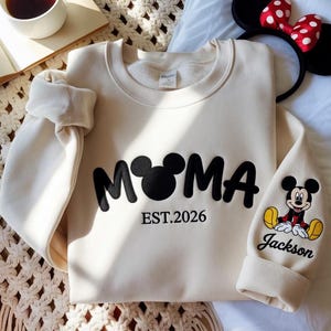 May include: Cream-colored sweatshirt with the word "MAMA" in black, featuring a Mickey Mouse head in place of the "O". The text "EST.2026" is below. The right sleeve has a Mickey Mouse graphic and the name "Jackson".