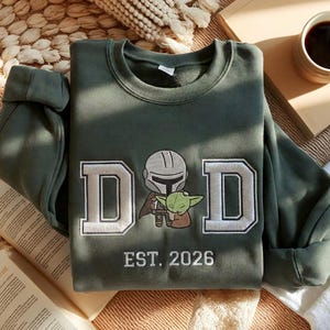 May include: Olive green sweatshirt with the word "DAD" in white embroidered letters, featuring an illustration of a Mandalorian and Grogu. The text "EST. 2026" is embroidered below. The sweatshirt is folded on a neutral surface.