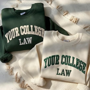 Könnte beinhalten: Zwei Sweatshirts, eines in Dunkelgrün und eines in Creme, werden gezeigt. Beide zeigen den Text "YOUR COLLEGE LAW" in einem College-Stil. Das grüne Sweatshirt hat weiße Buchstaben, während das cremefarbene grüne Buchstaben hat. Sie liegen auf einer strukturierten Oberfläche.