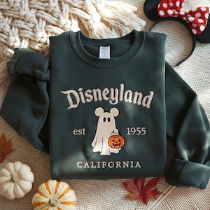 Pode incluir: Uma camisola de gola redonda verde-escura com o texto bordado Disneyland est 1955 California. O design central apresenta um fantasma branco com orelhas de rato a segurar um balde de ab&oacute;bora cor de laranja tipo lanterna.