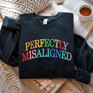 Op de afbeelding: Zwart sweatshirt met de geborduurde tekst "PERFECTLY MISALIGNED" in regenboogkleuren. Het sweatshirt is opgevouwen op een getextureerd oppervlak, met een boek en een kopje thee op de achtergrond. Een geschenkdoos is ook zichtbaar.