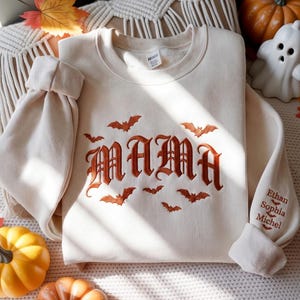 Könnte beinhalten: Ein cremefarbenes Sweatshirt mit Rundhalsausschnitt, auf dem das Wort MAMA in einer dunkelorangen gotischen Schrift gestickt ist, umgeben von kleinen gestickten Fledermäusen. Auf dem Ärmel stehen die Namen Ethan, Sophia und Michel in passender Schrift.