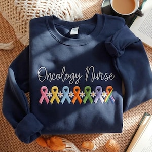 May include: A navy blue sweatshirt featuring the embroidered words "Oncology Nurse" in white script. Below are colourful awareness ribbons with daisy accents. The sweatshirt is folded on a textured surface.