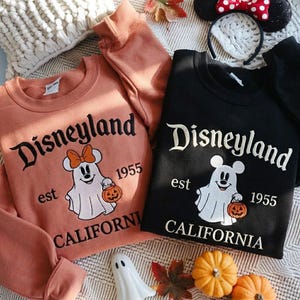 May include: Two crewneck sweatshirts, one in peach and one in black, featuring embroidered graphics of a ghost wearing mouse ears and holding a pumpkin. The text reads Disneyland, est 1955, and California. A headband with mouse ears is also visible.