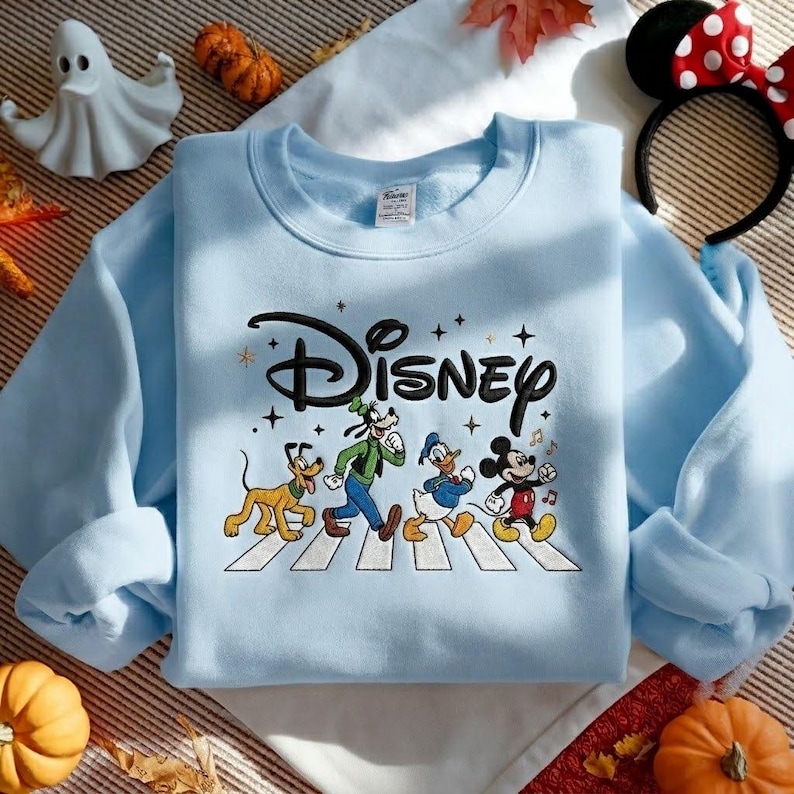 Peut inclure: Un sweat-shirt bleu clair &agrave; col rond arborant le logo Disney en noir au-dessus d'une illustration de Pluto, Dingo, Donald et Mickey marchant sur un passage pi&eacute;ton blanc. Le motif inclut de petites &eacute;toiles noires et des notes de musique.