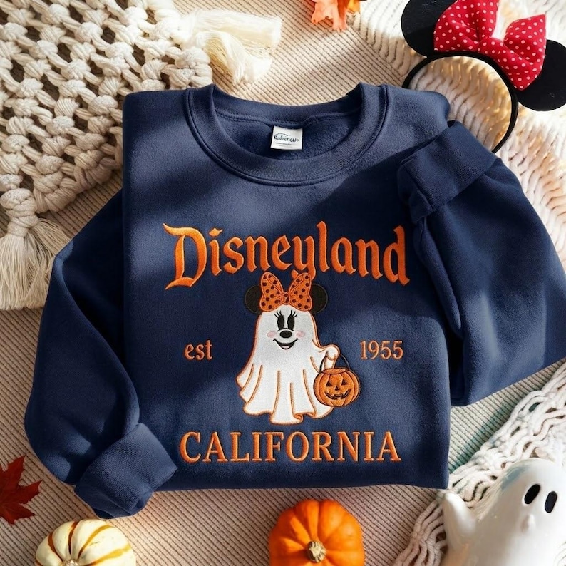 May include: A navy blue crewneck sweatshirt featuring orange text that reads Disneyland est 1955 CALIFORNIA. The center graphic shows a white ghost character wearing a polka dot bow and mouse ears, holding an orange jack-o'-lantern pumpkin bucket.