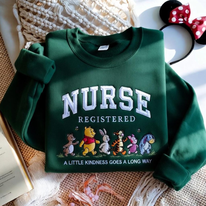 May include: A dark green sweatshirt with the word "NURSE" in white, collegiate-style lettering. Below, it says "REGISTERED" and features cartoon characters. The phrase "A LITTLE KINDNESS GOES A LONG WAY" is printed at the bottom.