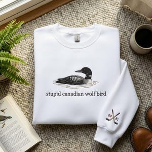 May include: White sweatshirt featuring a detailed illustration of a loon on the front, with the text "stupid canadian wolf bird" below. The sleeve has crossed hockey sticks with the numbers 24 and 81.