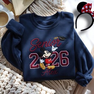May include: Navy blue sweatshirt with embroidered "Senior 2026" text, Mickey Mouse graphic, and personalized name. A graduation cap is also embroidered. A Minnie Mouse ear headband with a red polka dot bow is also visible.