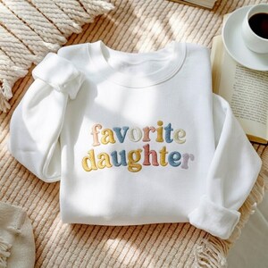 May include: White sweatshirt with the embroidered text "favorite daughter" in pastel colours. The sweatshirt is folded on a textured surface, with a book and a coffee cup in the background.