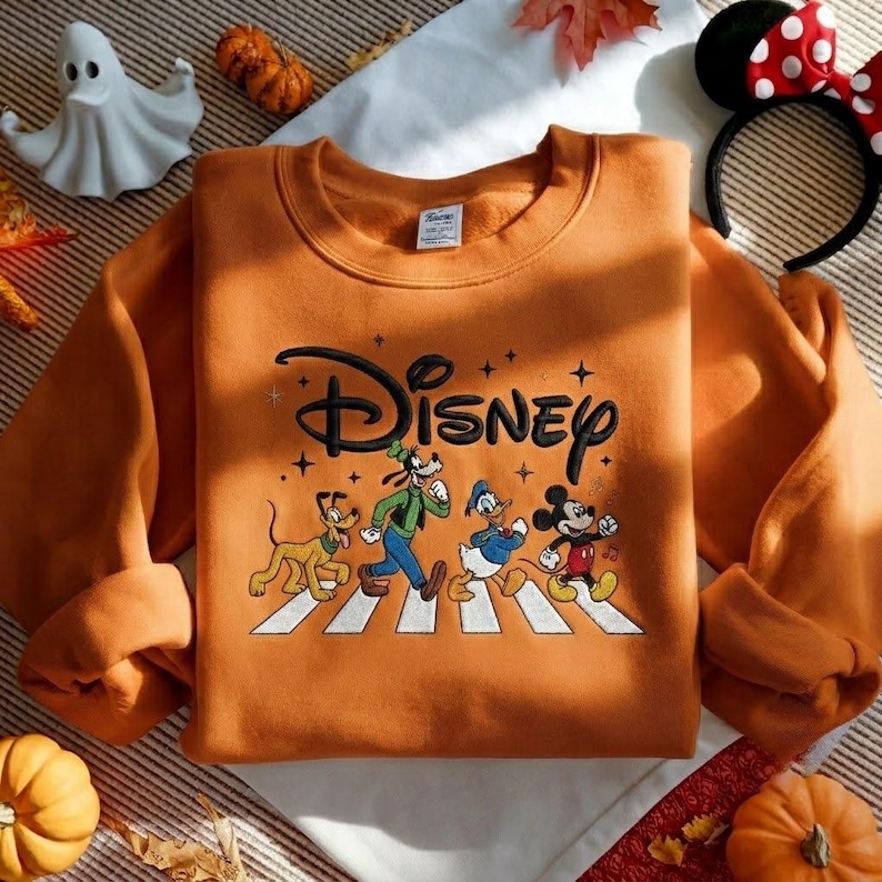 Peut inclure: Un sweat-shirt orange &agrave; col rond avec le logo Disney brod&eacute; en noir au-dessus d'un motif repr&eacute;sentant Pluto, Dingo, Donald et Mickey marchant sur un passage pi&eacute;ton blanc. Le design inclut des petites &eacute;toiles et des notes de musique.