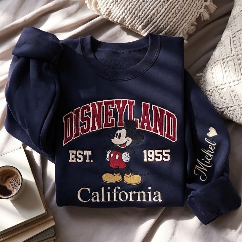 Puede incluir: Sudadera azul marino de Disneyland con Mickey Mouse. La sudadera muestra "Disneyland Est. 1955 California" en rojo y blanco. La manga tiene el nombre "Michel" en escritura blanca.