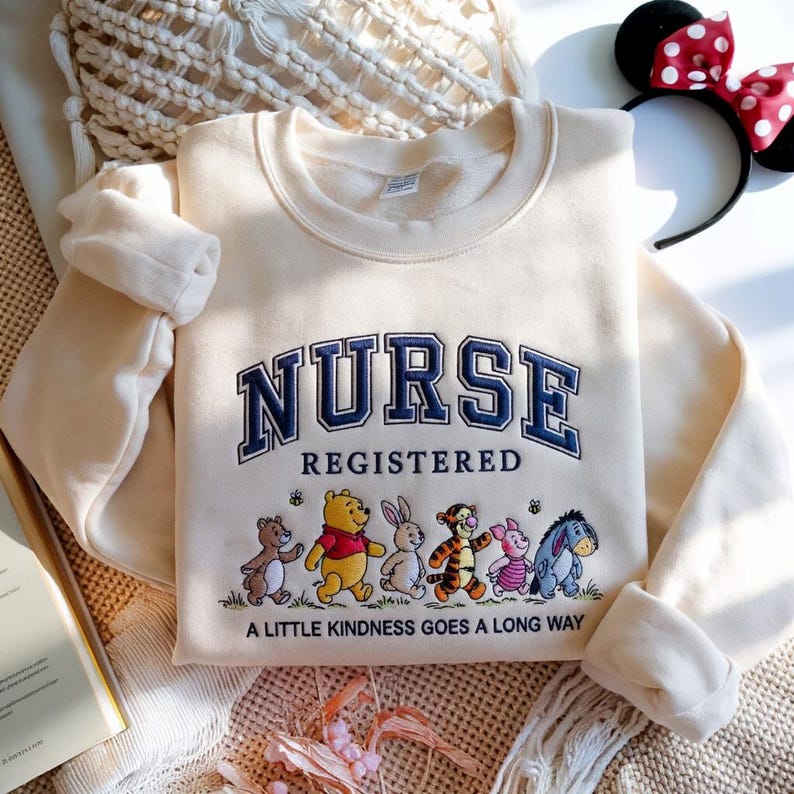 May include: Cream-colored sweatshirt with the word "NURSE" in navy blue collegiate lettering. Below, the word "REGISTERED" is displayed. The bottom of the sweatshirt features cartoon characters and the phrase "A LITTLE KINDNESS GOES A LONG WAY."