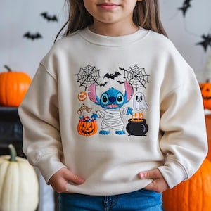 May include: Cream-colored sweatshirt featuring a Halloween-themed Stitch design. The design includes a mummy-wrapped Stitch, a jack-o'-lantern, a cauldron, a ghost, spiderwebs, and bats. The sweatshirt is a comfortable, casual style.