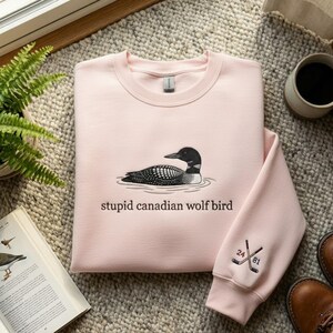 May include: A light pink sweatshirt featuring an embroidered loon design with the text "stupid canadian wolf bird". The sleeve cuff has crossed hockey sticks with the numbers "24" and "81".