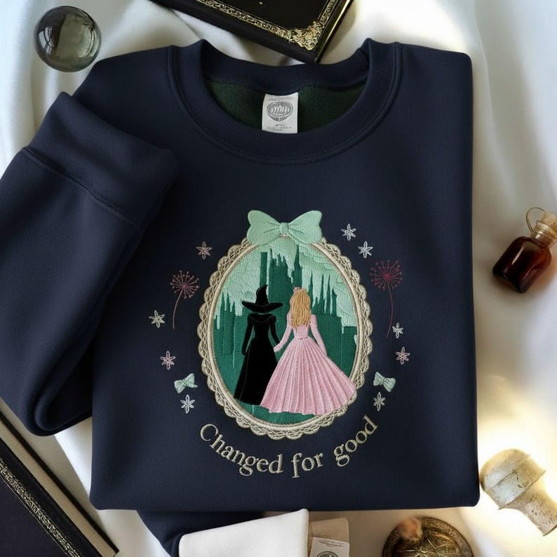 May include: Navy blue sweatshirt with an embroidered design featuring two figures in a frame with the text "Changed for good". The design includes a witch and a princess, with a green cityscape background and a mint green bow.