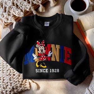 K&ouml;nnte beinhalten: Schwarzes Sweatshirt mit Minnie Mouse in einem rot-wei&szlig; gepunkteten Kleid und Schleife. Das Wort "MINNIE" ist in Blau, Gold und Rot gehalten. Der Text "SINCE 1928" steht darunter. Das Sweatshirt liegt auf einer neutralfarbenen Oberfl&auml;che.