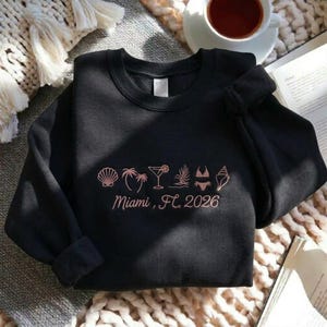Peut inclure: Sweat-shirt noir &agrave; col rond avec un motif corail repr&eacute;sentant Miami, FL 2026. Le motif comprend des &eacute;l&eacute;ments de plage tels que des coquillages, des palmiers, un cocktail et un bikini.
