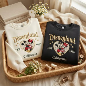 May include: Two crewneck sweatshirts, one cream and one black, featuring the Disneyland logo with Mickey and Minnie Mouse in a heart. The sweatshirts have gold embroidery and the text "EST 1955 California". They are displayed in a woven basket.