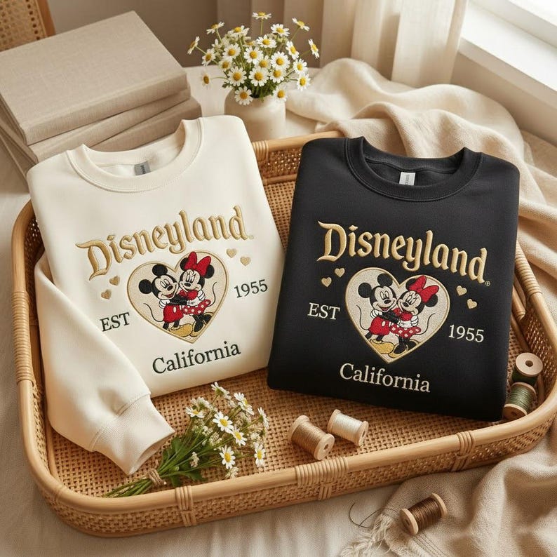 May include: Two crewneck sweatshirts, one cream and one black, featuring the Disneyland logo with Mickey and Minnie Mouse in a heart. The sweatshirts have gold embroidery and the text "EST 1955 California". They are displayed in a woven basket.