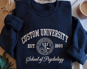 May include: A navy blue crewneck sweatshirt featuring white embroidered text that reads CUSTOM UNIVERSITY EST. 1889 and School of Psychology. The center includes a circular emblem with a Greek psi symbol, a book, and a brain illustration.