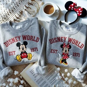 May include: Two gray sweatshirts with the text "DISNEY WORLD EST 1971" in red embroidery. One sweatshirt features Mickey Mouse, the other Minnie Mouse. A black headband with a red and white polka dot bow is also visible.