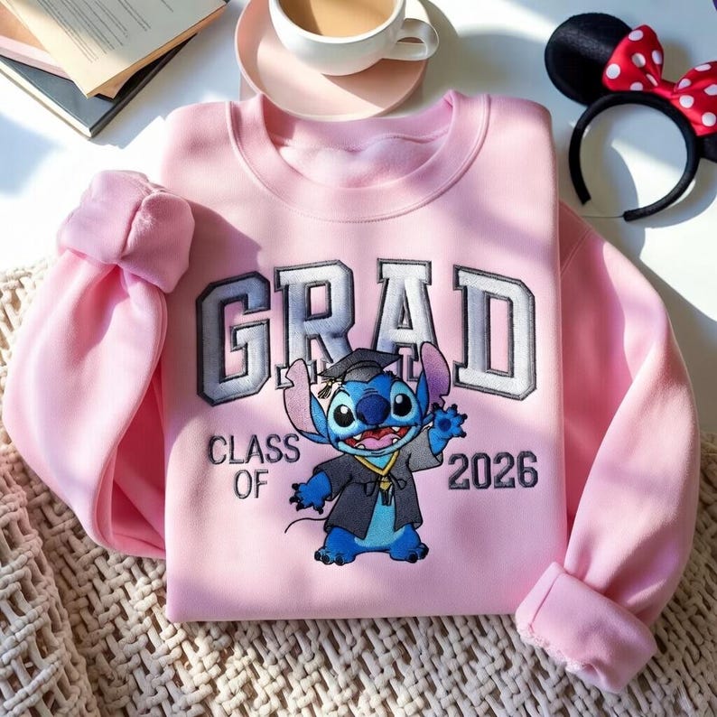 Puede incluir: Sudadera rosa claro con la palabra "GRAD" en letras grises y blancas, y el texto "CLASS OF 2026". Presenta un bordado del personaje Stitch con toga de graduaci&oacute;n.