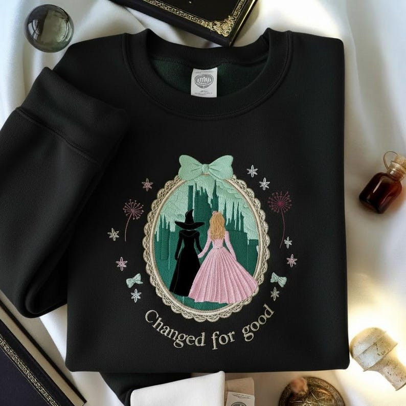 May include: Black sweatshirt with an embroidered design featuring two figures in front of a green castle. One figure wears a black dress and hat, the other a pink dress. The text "Changed for good" is below the design.