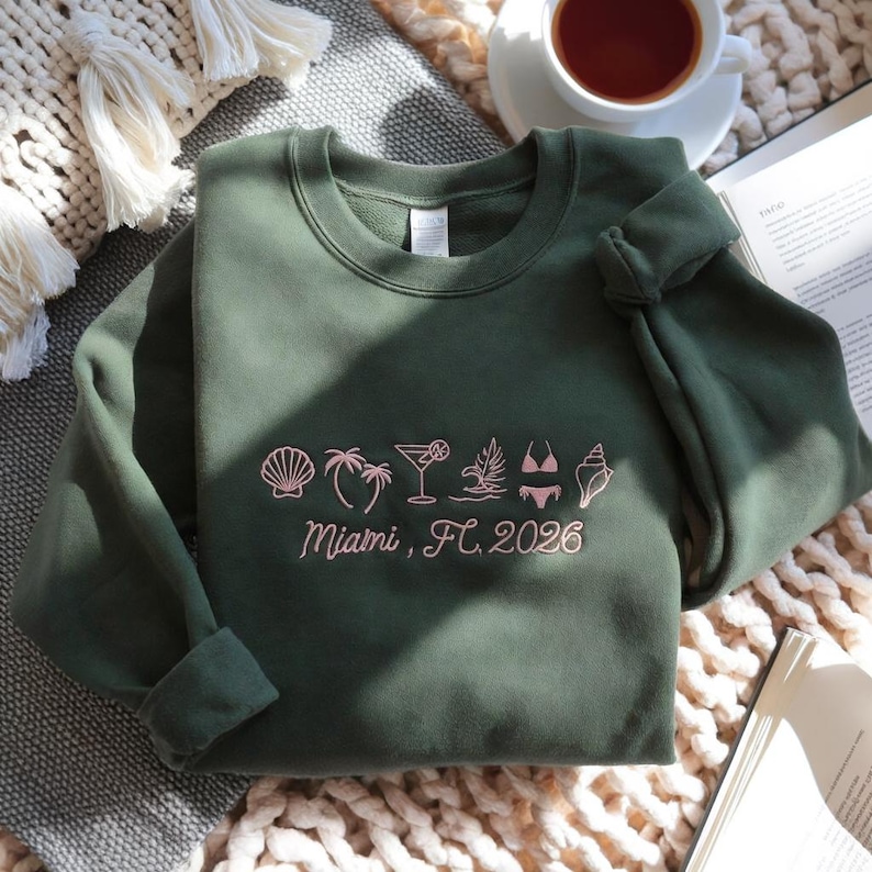 Peut inclure: Un sweat-shirt vert olive avec des ic&ocirc;nes roses brod&eacute;es d'un coquillage, de palmiers, d'un cocktail et d'un bikini. Le texte "Miami, FL 2026" est brod&eacute; en dessous des ic&ocirc;nes.