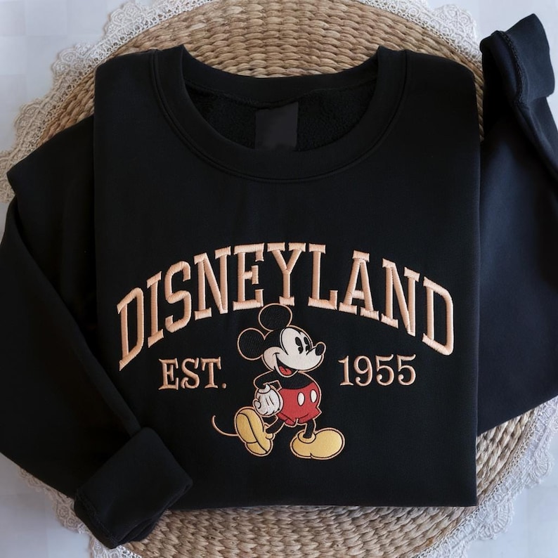 May include: Black sweatshirt with tan "Disneyland" text, "EST. 1955", and a Mickey Mouse graphic. The sweatshirt is folded on a woven surface.