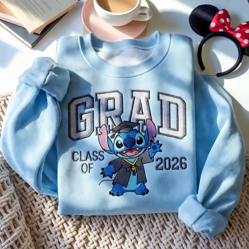 Puede incluir: Sudadera azul claro con un dise&ntilde;o tem&aacute;tico de graduaci&oacute;n. La palabra "GRAD" est&aacute; bordada en gris y blanco, con "CLASS OF 2026" debajo. Un personaje de dibujos animados con toga de graduaci&oacute;n est&aacute; en el centro.