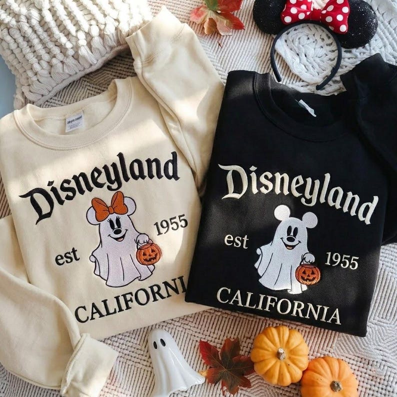 May include: Two crewneck sweatshirts, one cream and one black, featuring embroidered graphics of a ghost wearing mouse ears and holding a pumpkin bucket. The text reads Disneyland est 1955 California. The cream sweatshirt ghost wears an orange bow.