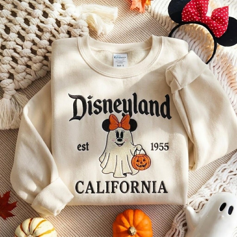 May include: A cream-colored crewneck sweatshirt featuring black embroidered text that reads Disneyland est 1955 California. The center design shows an embroidered ghost wearing a Minnie Mouse bow and holding an orange jack-o'-lantern pumpkin.