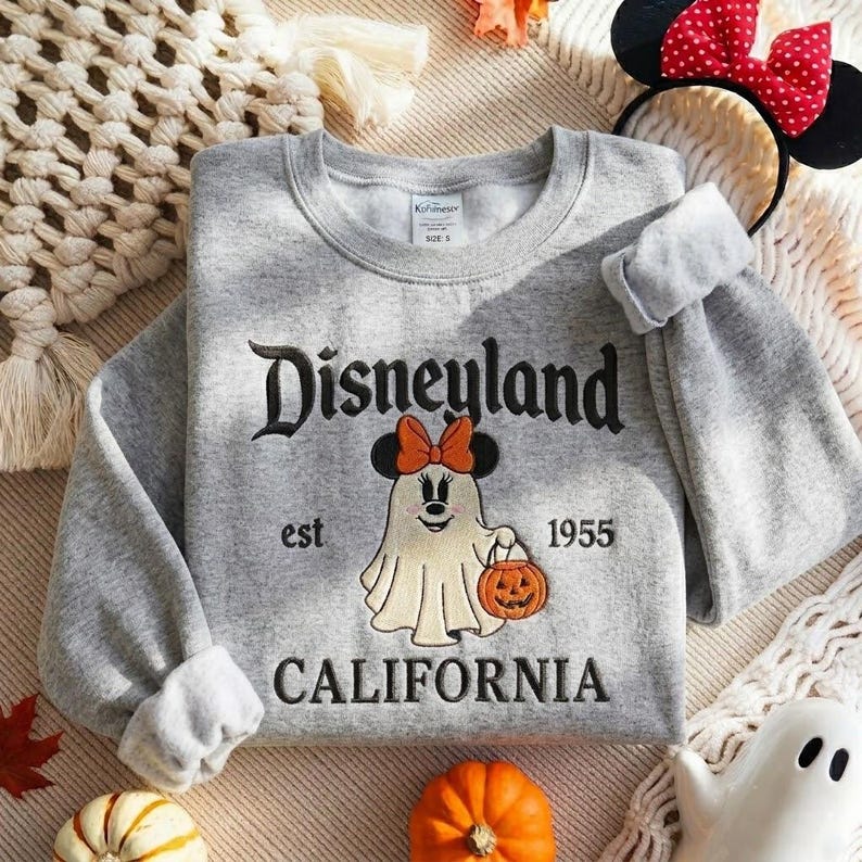 May include: A heather gray crewneck sweatshirt featuring embroidered black text that reads Disneyland est 1955 CALIFORNIA. The center graphic depicts a white ghost character wearing an orange bow and holding a small orange jack-o'-lantern.