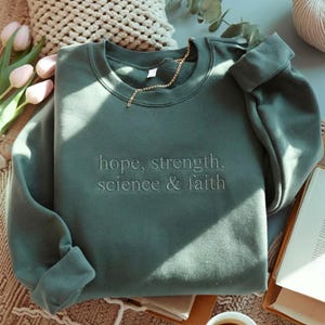 Peut inclure: Un sweat-shirt vert sauge avec les mots "hope, strength, science & faith" brodés sur le devant. Une chaîne dorée est drapée autour de l'encolure. Le sweat-shirt est plié sur une table avec un livre et une tasse de café.