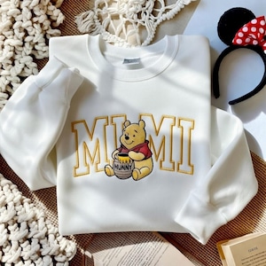 以下が含まれることがあります： 金色の文字で「MIMI」と書かれた白いスウェットシャツと、くまのプーさんのイラスト。漫画のキャラクターは蜂蜜の壺を持っています。赤と白の水玉模様のリボンが付いた黒いヘアバンドが背景にあります。