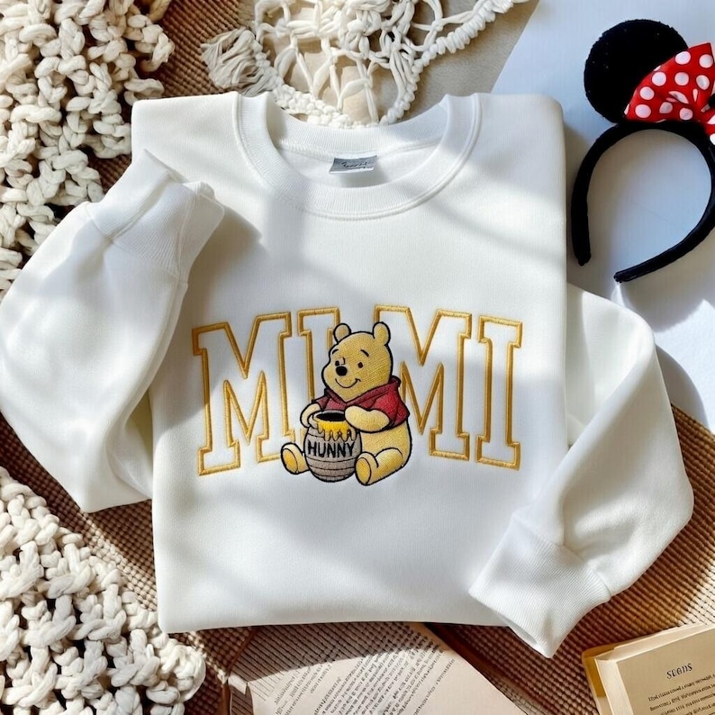 以下が含まれることがあります： 金色の文字で「MIMI」と書かれた白いスウェットシャツと、くまのプーさんのイラスト。漫画のキャラクターは蜂蜜の壺を持っています。赤と白の水玉模様のリボンが付いた黒いヘアバンドが背景にあります。