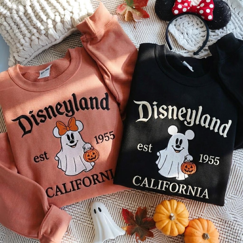 May include: Two crewneck sweatshirts, one in peach and one in black, featuring embroidered graphics of a ghost wearing mouse ears and holding a pumpkin. The text reads Disneyland, est 1955, and California. A headband with mouse ears is also visible.