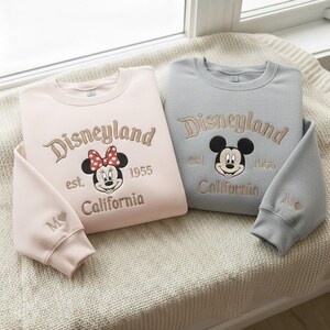 Puede incluir: Dos sudaderas de cuello redondo, una rosa claro y otra gris, con texto bordado "Disneyland", Mickey y Minnie Mouse, y texto "California". Las mangas tienen una inicial y un coraz&oacute;n bordados.