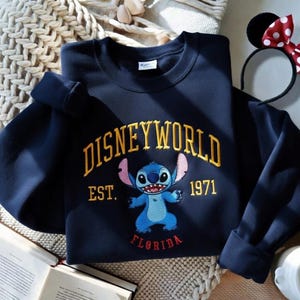 May include: Navy blue sweatshirt with "DISNEYWORLD" in gold lettering and a blue Stitch character graphic. The sweatshirt also features "EST. 1971" and "FLORIDA" in gold and red lettering. A Minnie Mouse ear headband is visible.